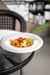 Tagliatelle pasta with chicken, mushrooms, and cherry tomatoes served on a white plate outdoors. Casual dining setting with glass table and wicker chair in natural light.
