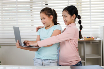 Two girls are standing with laptops in a classroom or study room with a large window. One is...