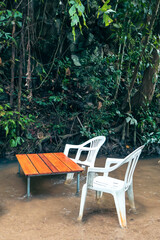 Obraz premium Outdoor seating area with white plastic chairs, rustic wooden table, surrounded by lush greenery, perfect for relaxation and casual gatherings.