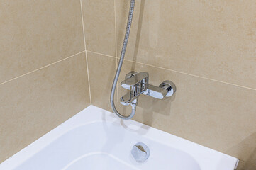 Close-up of a bathtub with a chrome faucet and shower hose against tiled beige walls