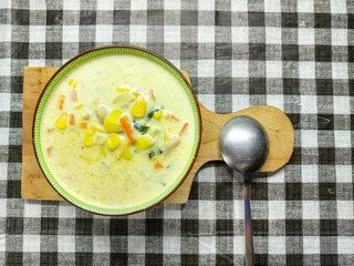 A delicious bowl of homemade cream soup with mixed vegetables like corn and carrots, viewed from above on a checkered placemat