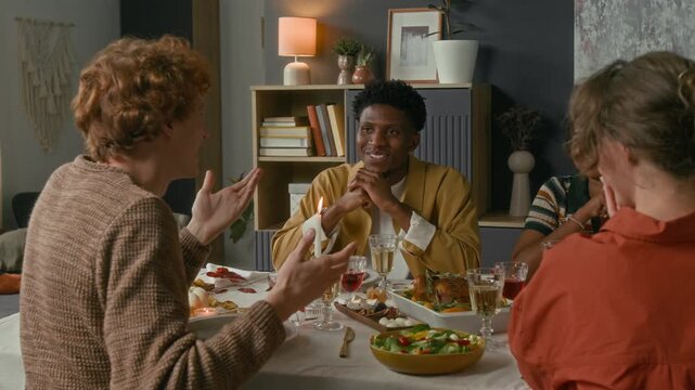 Joyful Caucasian young man actively gesticulating telling fun story to three positive diverse friends while having holiday lunch on thanksgiving day