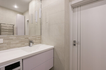 Modern bathroom features light beige tiling, a white vanity with a sink, a sleek faucet, and a door. The space is clean and well-lit