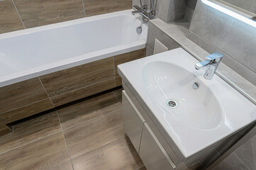 Bright bathroom featuring a white sink with chrome faucet, alongside a bathtub with wood-look tiling. Gray wall tiling complements the clean, modern aesthetic