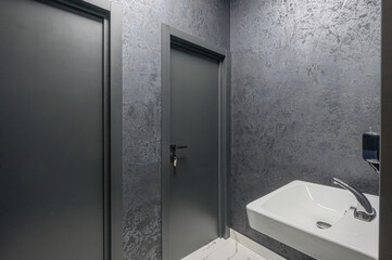 modern bathroom interior features textured grey walls, two dark grey doors, a white wall-mounted sink with a chrome faucet, and white marble-patterned flooring