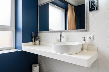 A chic bathroom featuring a modern, round white sink, a clean white countertop, a framed mirror, and stylish white herringbone tiles against a deep blue wall