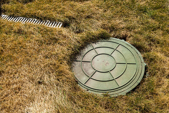 Overhead view of a green septic tank lid nestled in dry, golden grass next to a white drainage grate