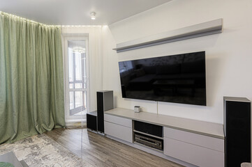 Bright living room with a large TV mounted above a gray cabinet. Green curtains flank a window. Speakers are positioned on either side of the media center. Wood flooring adds warmth