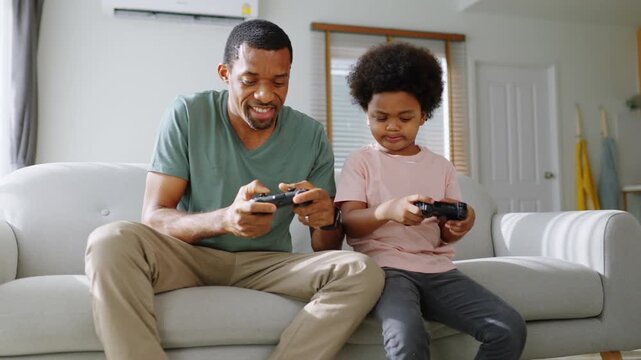 Joyful African father and son sitting on sofa playing console games together, laughing and bonding during fun family time at home.