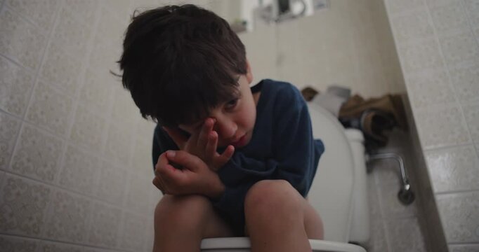 Young boy sitting on toilet in morning after waking up, sleepy child with hand on face using bathroom, candid everyday routine of childhood hygiene at home
