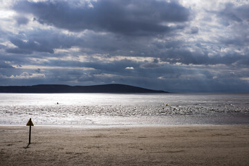 Dramatic clouds over a beach at low tide. A warning sign stands on the sand, cautioning about the mudflats.
