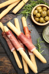 Delicious grissini sticks with prosciutto, peppercorns, green olives and rosemary on wooden table, flat lay