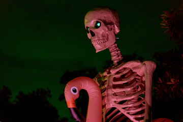 A large skeleton figure with glowing eyes stands at night holding a pink flamingo prop. Festive Halloween yard display with dramatic green sky and colorful lighting.