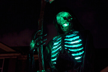 Hooded skeleton reaper holds a scythe with bright green ribs and eyes at night. Spooky Halloween...