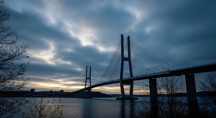 Obraz premium A dramatic cable-stayed bridge stretches across a calm body of water at dawn, silhouetted against a brooding sky filled with dramatic clouds.