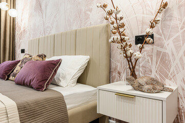 A stylish bedroom features a beige bed with pillows, a white nightstand with decor, and patterned...