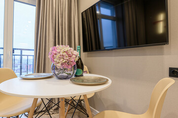 Cozy apartment scene: white table with flowers, champagne, plates, and chairs beneath a wall-mounted TV