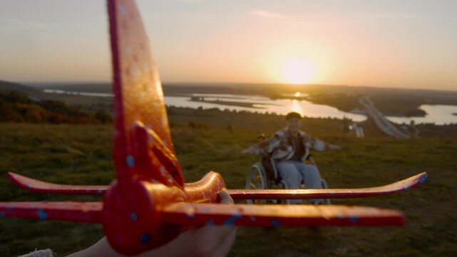 Open field is flooded with warm evening light, parent makes plane soar into air, and the child in wheelchair raises his arms, imitating feeling of floating in sky dreaming of flying.