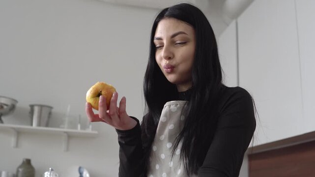 young woman in apron eats apple in the kitchen, girl bites the fruit and relaxes at home