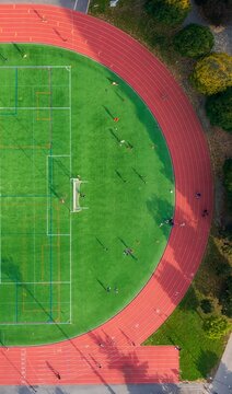 Aerial view of a vibrant green sports field bisected by stark white lines, bordered by a russet running track, a hive of activity under the open sky, Brooklyn, New York, United States.