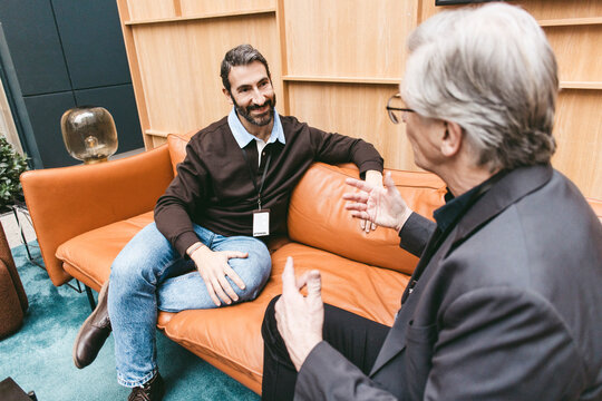 Smiling businessman listening to senior colleague discussing while sitting on sofa at networking event in convention cen