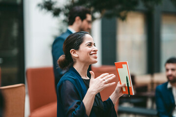 Smiling female business expert holding book gesturing while talking in convention center