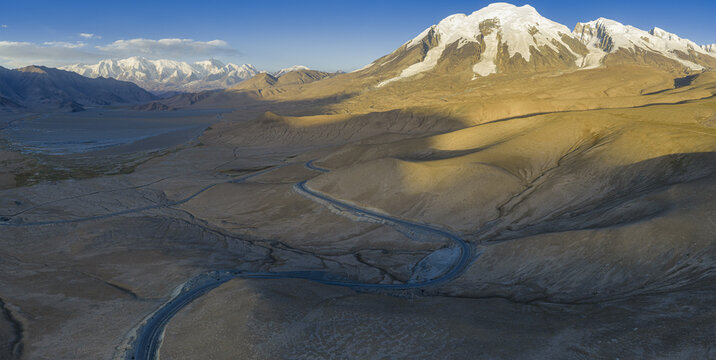 Aerial view of a winding road cutting through the rugged terrain towards the snow-capped Muztagh Ata, a stark contrast under the vast sky, Muztagh Glacier, China.
