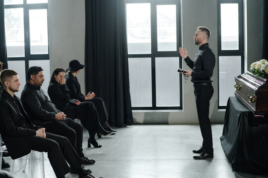 Caucasian man standing and speaking to group of men and women sitting in row at funeral ceremony near closed casket in modern room 