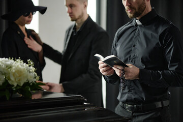 Caucasian middle aged man in clerical clothing holding prayer book and rosary standing near coffin while Caucasian young adult man comforting woman in background