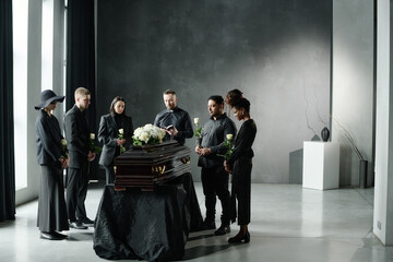 Group of middle aged and young adult men and women standing around closed casket holding roses...