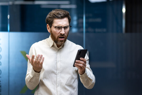 A stressed young businessman is standing in the office and looking at his mobile phone in frustration, spreading his arms