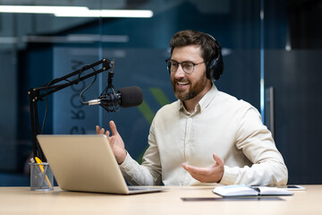A young man wearing headphones sits in an office in front of a microphone and talks into a laptop camera, gesturing with his hands