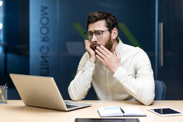 A tired and overworked young man sits in the office at a desk with a laptop and yawns