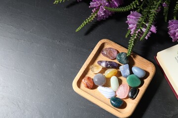 Different beautiful gemstones, book and flowers on black table, flat lay. Space for text