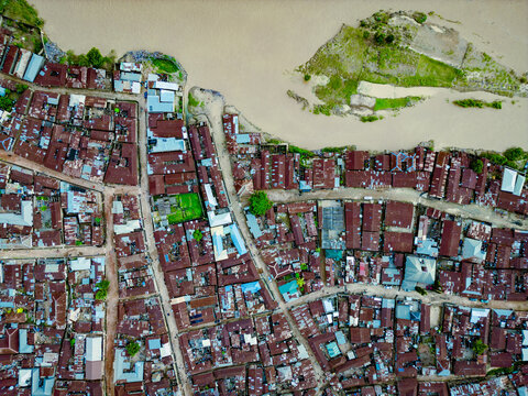 Aerial view of a densely packed town with rusty-brown rooftops contrasting against the muddy river and green vegetation, Kabala Costain, Kaduna, Nigeria.