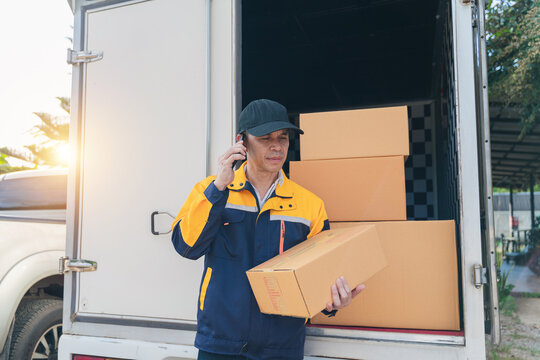 Delivery professional talking on mobile phone while holding package next to parked vehicle with shipment boxes in background during sunset