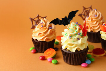 Delicious cupcakes with bat topper, red leaves, sprinkles and candies on orange background, closeup with space for text. Halloween celebration