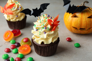 Delicious cupcakes with bat topper, branches with red leaves, sprinkles, candies and pumpkin on grey textured table, closeup. Halloween celebration