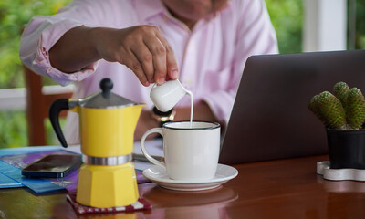 Business human pouring milk into hot coffee cup while working on laptop at home office with moka pot and cactus plant on wooden desk, cozy morning routine.