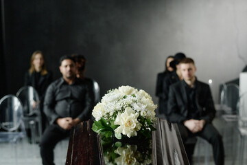 Group of middle aged and young adult Caucasian and Black men and women sitting in rows attending funeral service, floral arrangement on closed casket in foreground, somber expressions