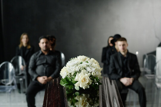 Group of middle aged and young adult Caucasian and Black men and women sitting in rows attending funeral service, floral arrangement on closed casket in foreground, somber expressions