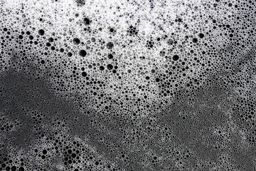 White foam bubbles spread across dark liquid surface, creating dynamic and effervescent abstract pattern. Varying sizes of delicate bubbles convey sense of movement and freshness, suggesting clean
