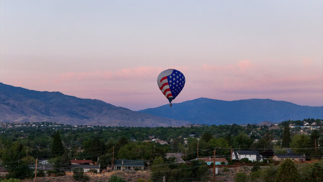 Aerial view of a vibrant hot air balloon adorned with the American flag ascends gracefully over the cityscape, set against a backdrop of dusky mountains, Reno, Nevada, United States.