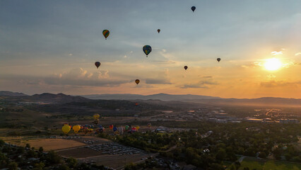 Aerial view of hot air balloons ascending into the golden sunrise over the city and mountains, their vibrant colors contrasting against the soft hues of the sky, Reno, Nevada, United States.