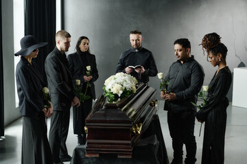 Group of middle aged and young adult multiethnic men and women standing around coffin holding...