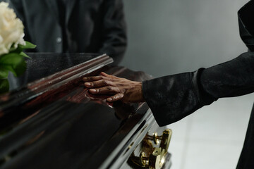Black woman touching closed coffin during funeral ceremony, hand resting gently on polished wood surface, expressing grief and loss, floral arrangement partially visible nearby