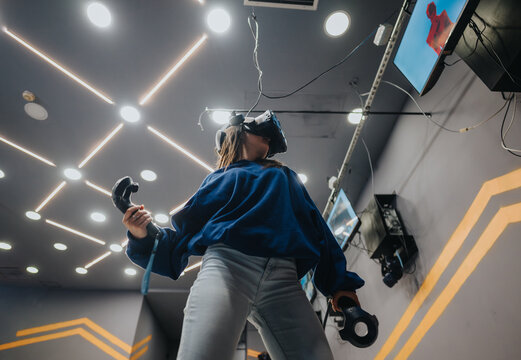 A woman actively immersed in virtual reality game play, holding controllers and wearing a VR headset. The setting features futuristic lighting, emphasizing advanced digital gaming experiences.