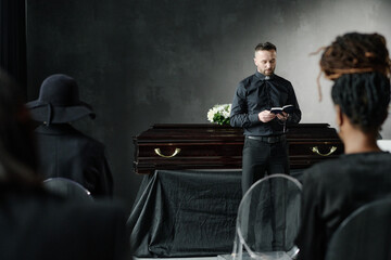 Caucasian young adult man reading from book standing in front of closed coffin during funeral ceremony with Black women sitting in foreground listening attentively