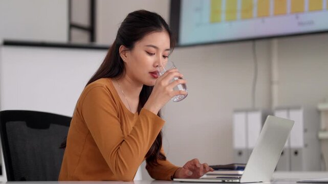 Asian adult businesswoman use laptop computer stay hydrated drink water modern office workspace productivity