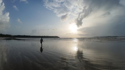 Golden Hour Solitude: A Person Walking on a Reflective Sandy Beach as the Sun Sets Dramatically Behind the Horizon and Breaks Through the Clouds